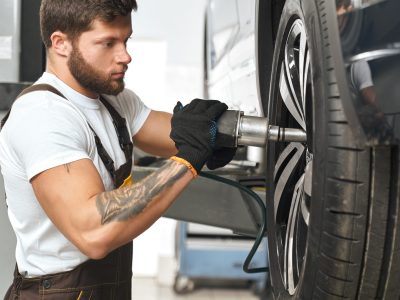 Bearded, brutal mechanic fixing hubcap of auto wheel, using equipment. Muscular man in white t shirt with tattoo on hand wearing in black gloves, working in autoservice.